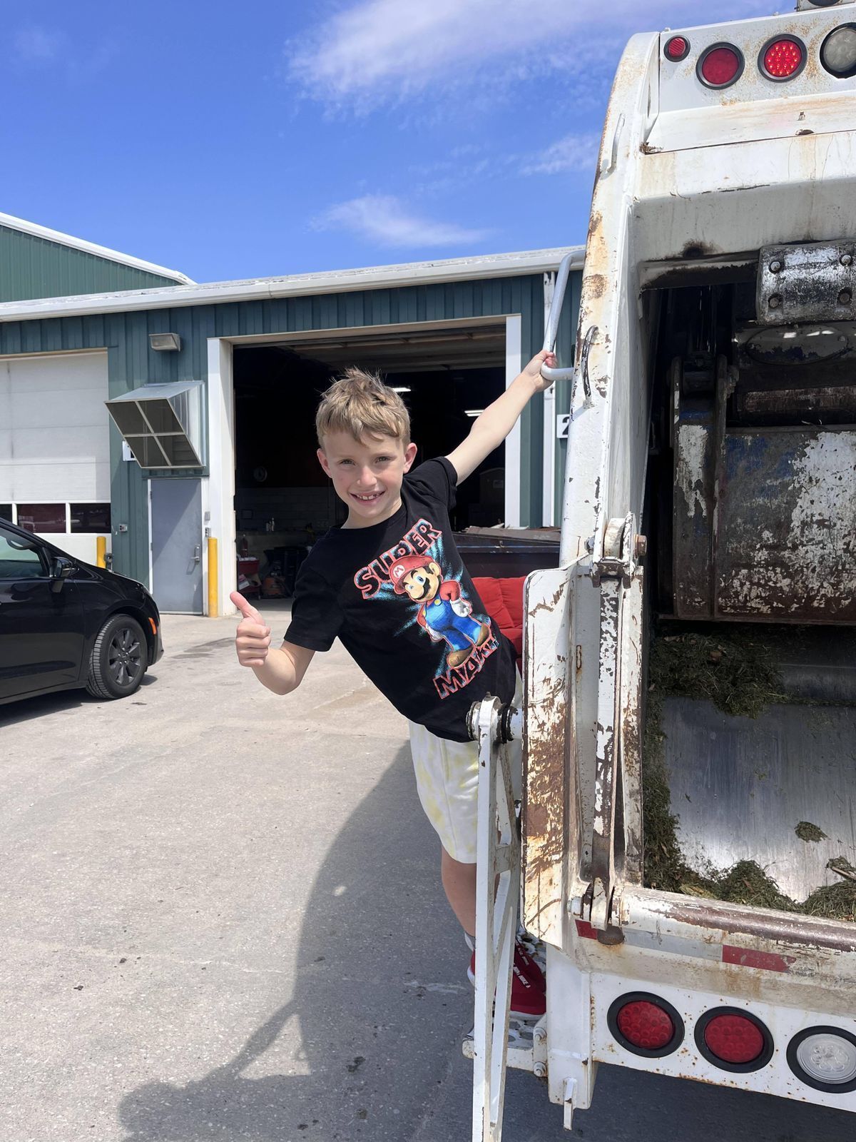 A young boy is standing next to a garbage truck and giving a thumbs up.
