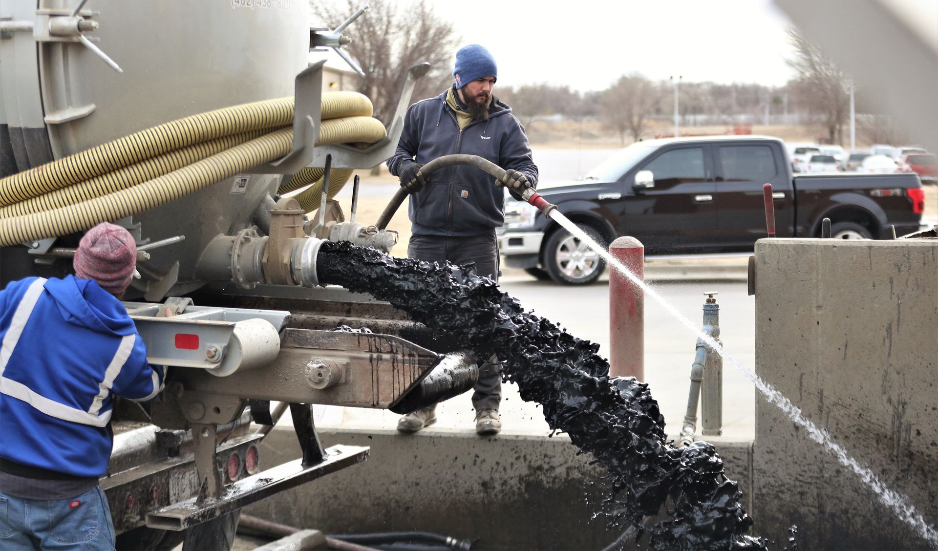 A man in a blue jacket is standing next to a truck with a hose attached to it. A man in a blue jacket is standing next to a truck with a hose attached to it.