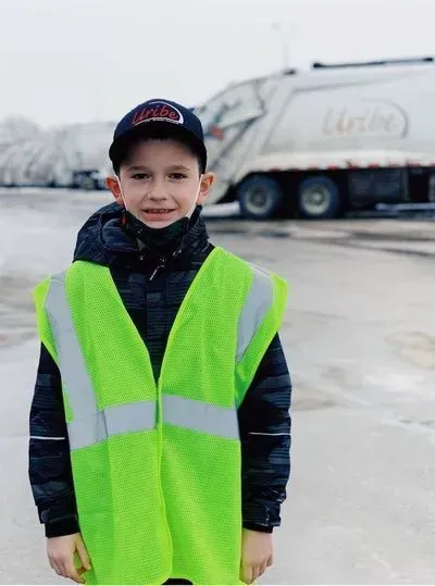 A young boy wearing a yellow vest and hat stands in front of a garbage truck