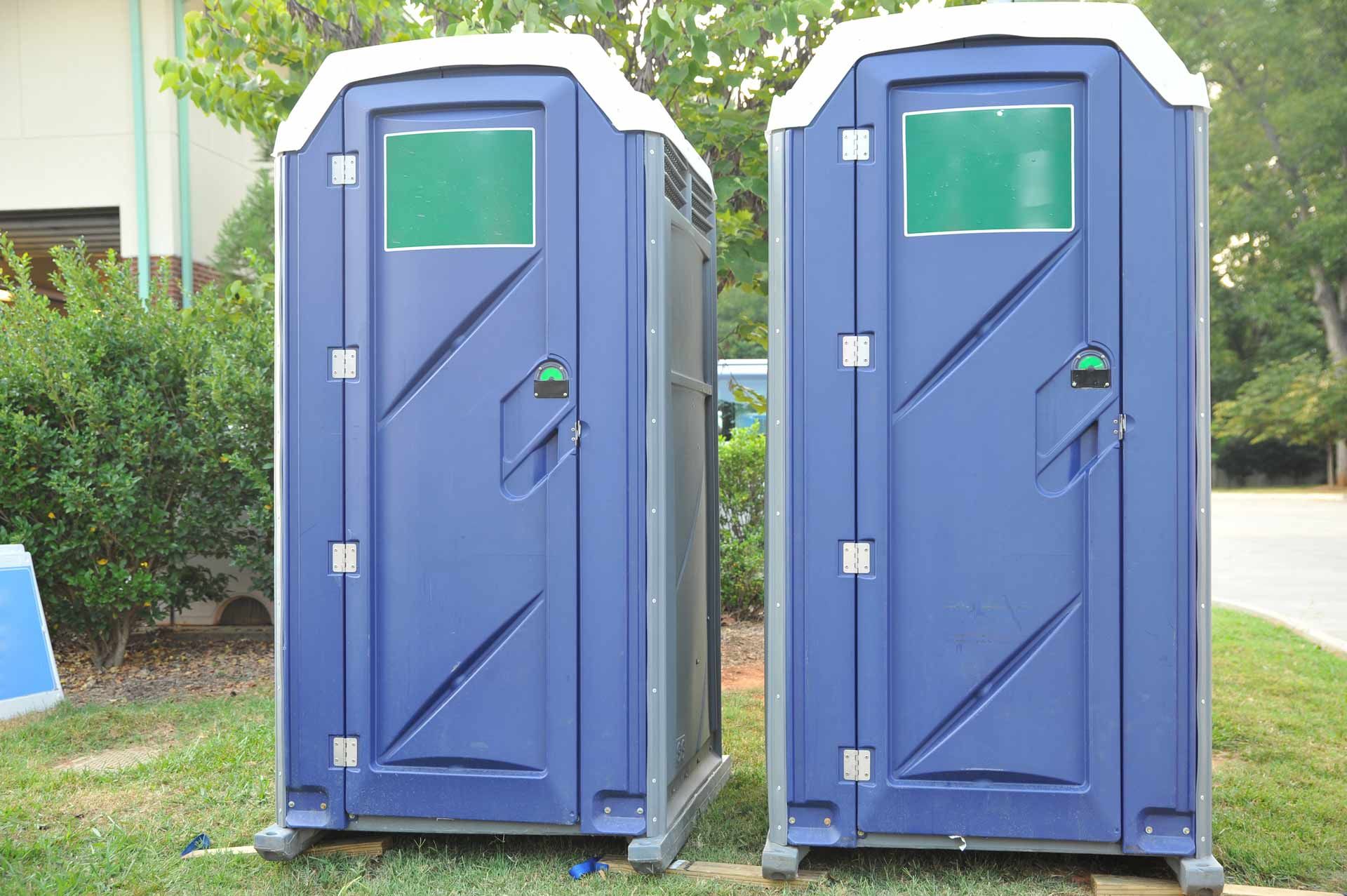 Two portable toilets with navy blue doors stand side by side on a yard, with bushes close-by. Two portable toilets with navy blue doors stand side by side on a yard, with bushes close-by.