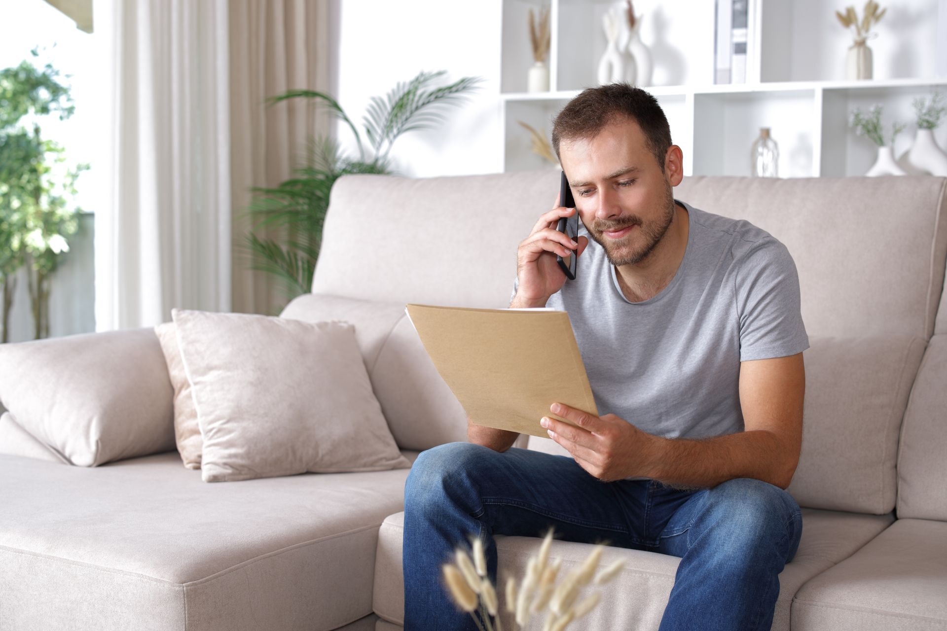 Man on phone, looking at paper. Sitting on beige couch in a living room.