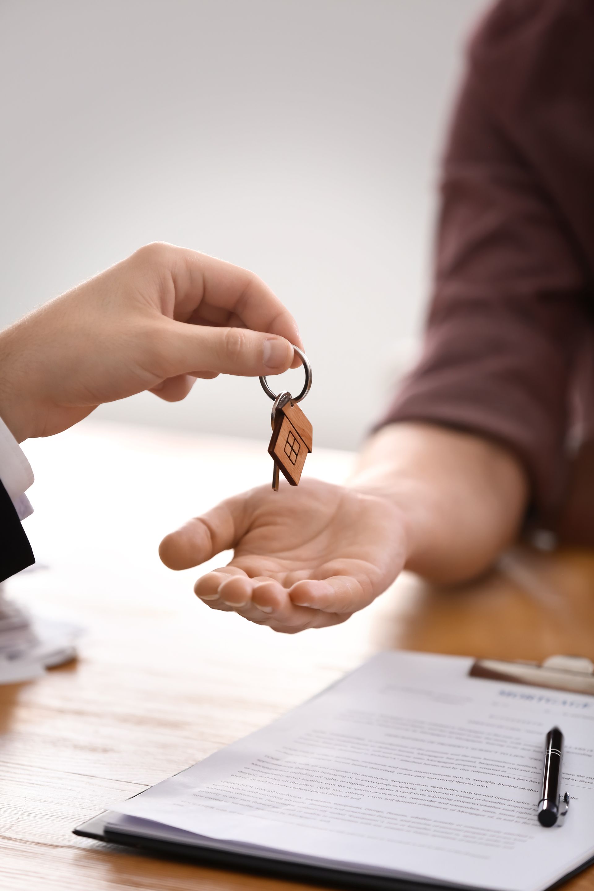 Person handing keys with a wooden keychain to another over a document on a table.