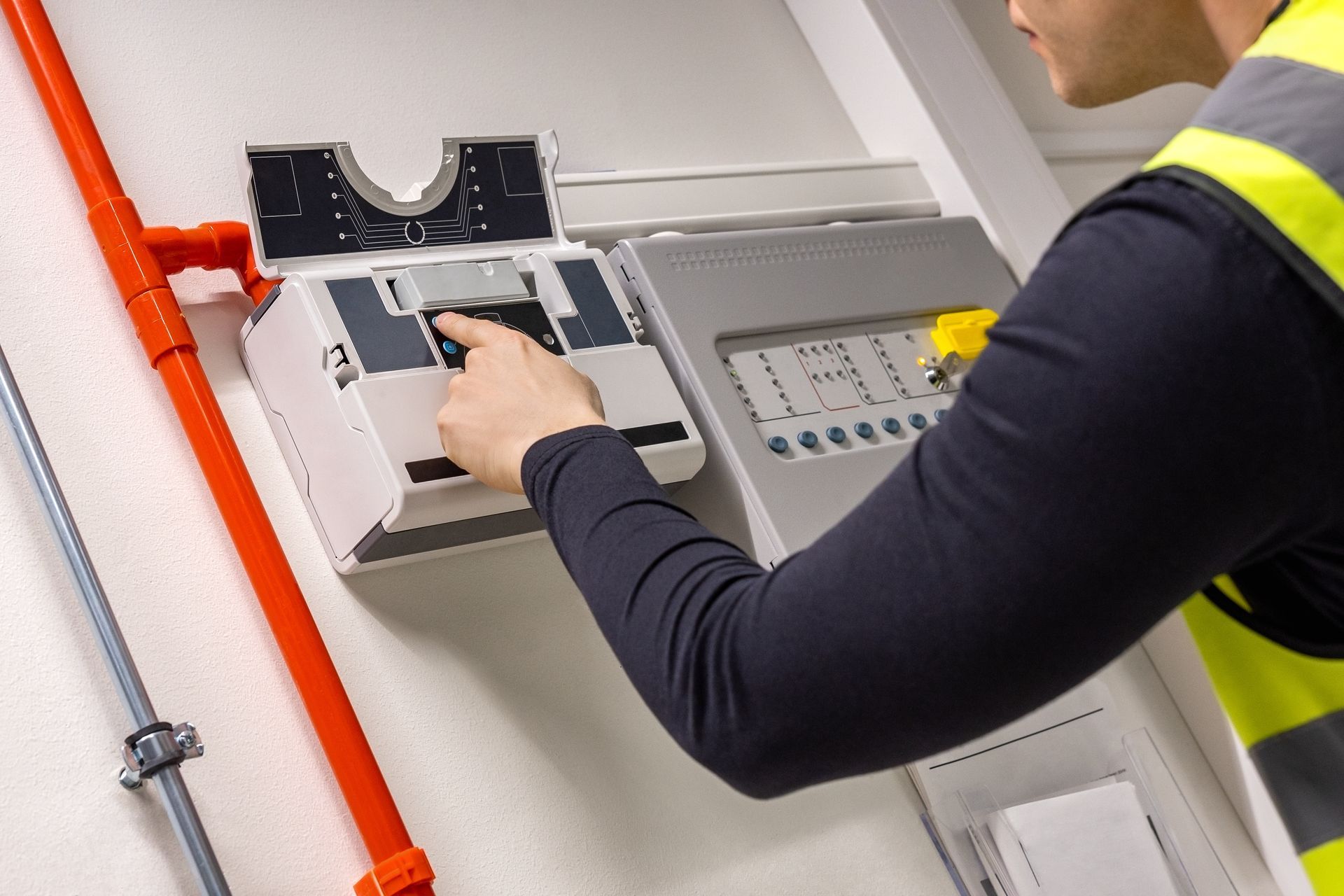 Person in a safety vest presses a button on a fire alarm control panel near orange pipes.
