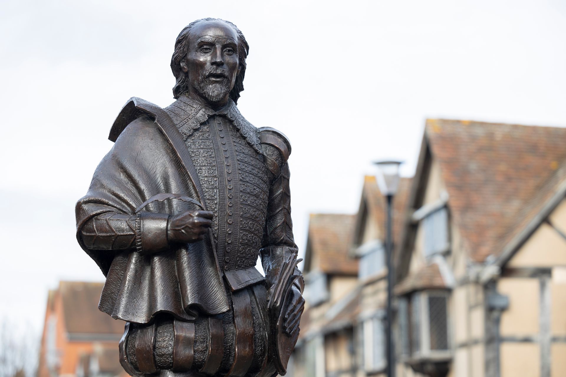 Statue of Shakespeare in Stratford-upon-Avon, wearing period clothing, in front of Tudor-style buildings.