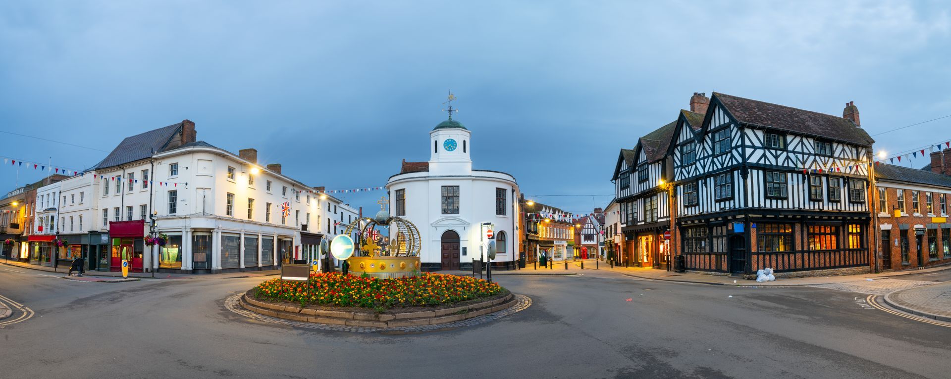 Town square with buildings, a white clock tower, and a roundabout with flowers. Cloudy sky at dusk.