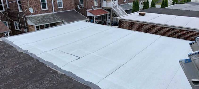 View of a large white flat roof. Brick buildings in the background. The sky is overcast.