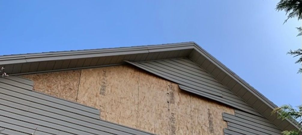 The side of a house with missing siding, showing exposed plywood, under a clear blue sky.