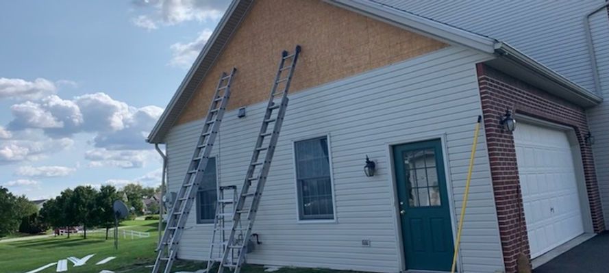 Two ladders lean against a house with exposed wood under the roof. The sky is blue and a green yard is visible.