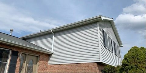 Exterior view of a house with grey siding, brick foundation, and dark shutters. The sky is partly cloudy.
