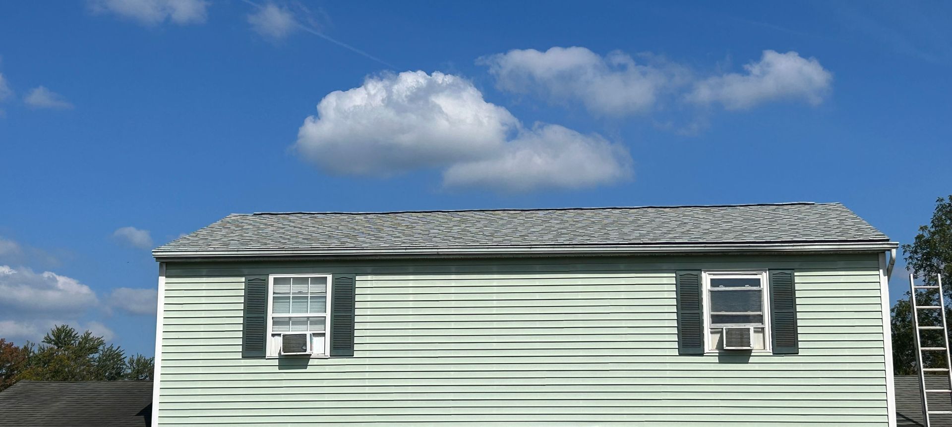A light green house with gray roof, black shutters, and two windows sits under a blue sky with white clouds.