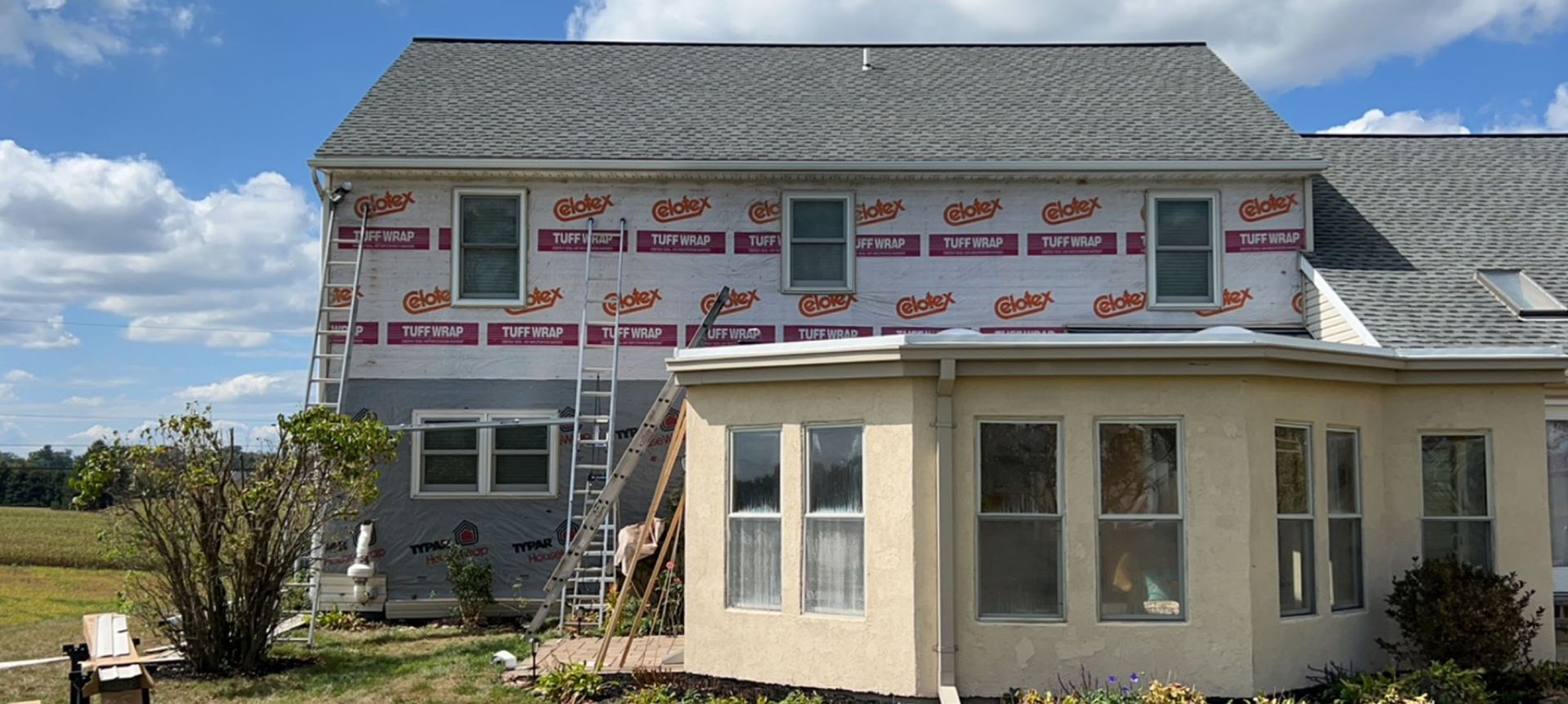 A two-story house with under-construction siding and a sunroom, set against a blue sky.