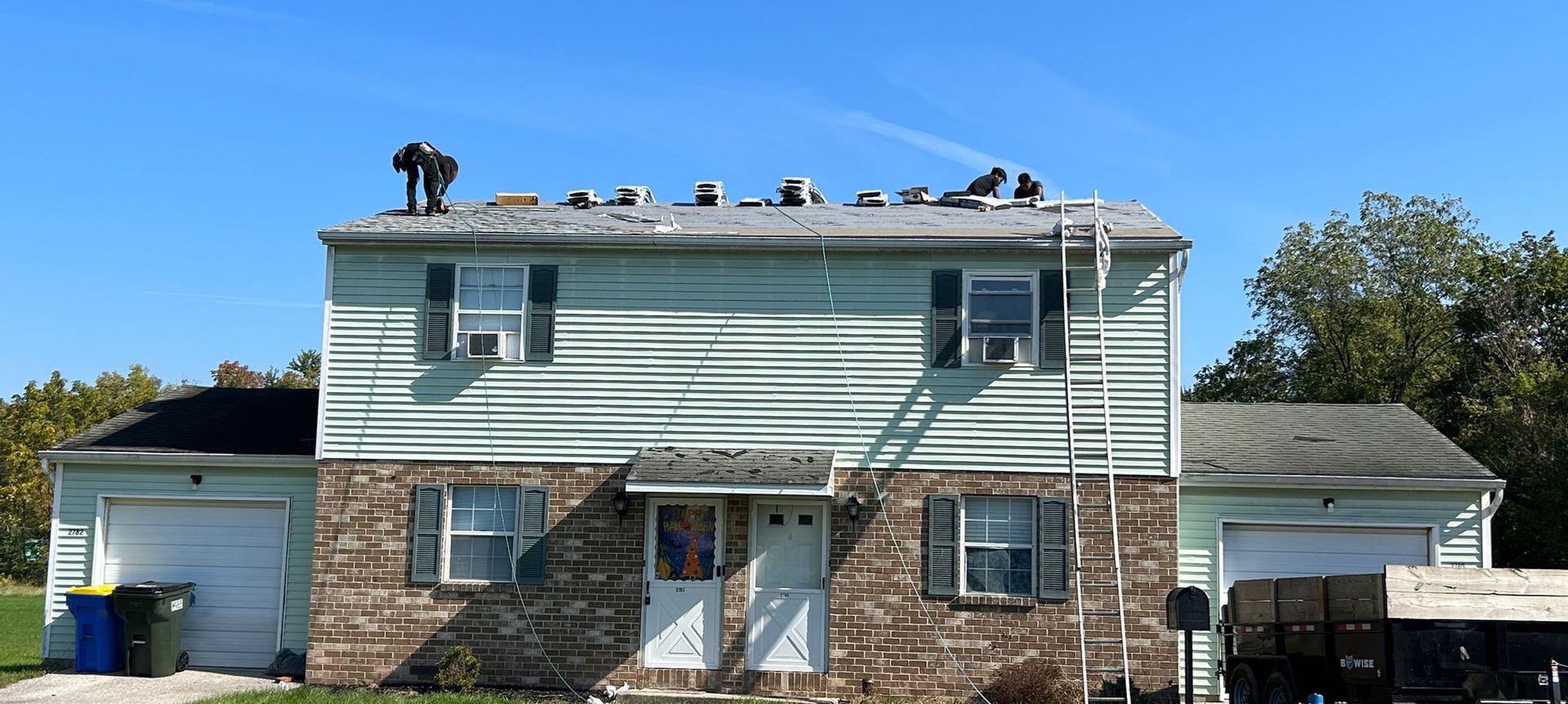 Workers on a two-story building's roof, repairing shingles. A ladder is present. Blue sky.