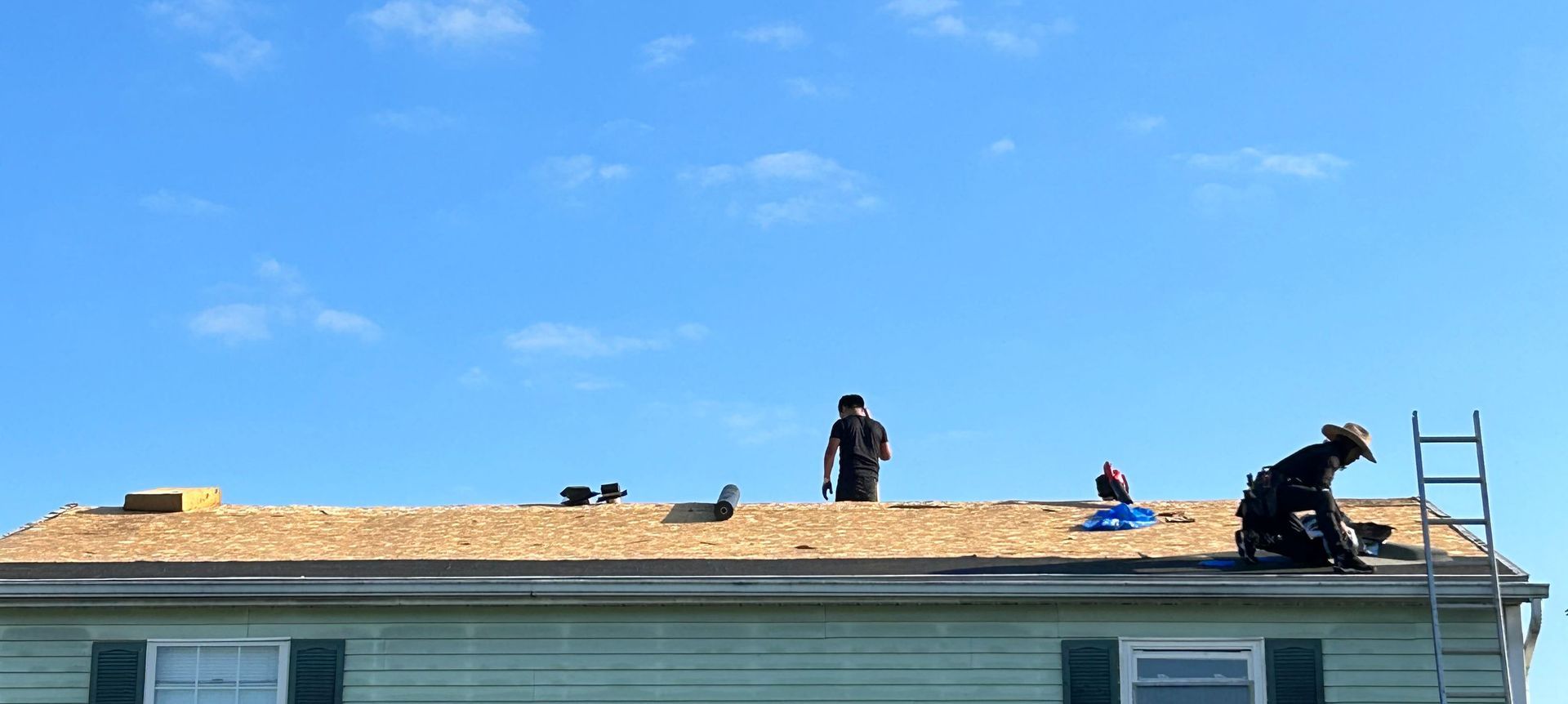 Two roofers on a rooftop under a bright blue sky, one near a ladder, the other holding a tool.