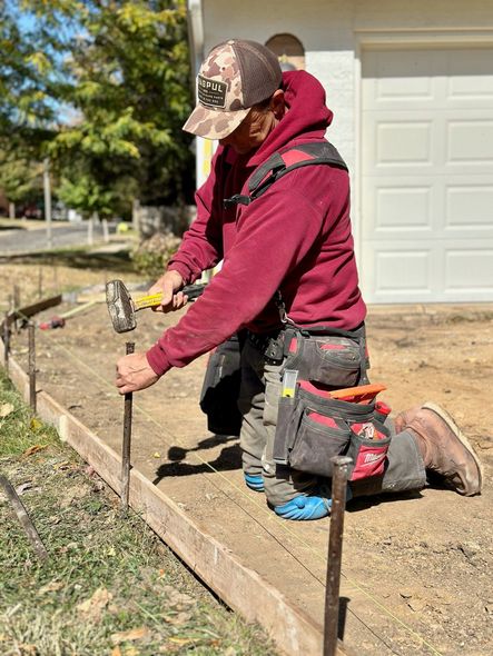 A worker in a maroon hoodie and camo cap kneels, using a mallet to drive a metal stake into the ground for a wood form.
