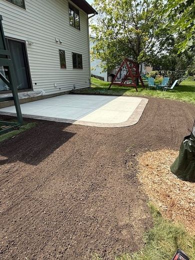 A new concrete patio with a brick border sits next to a light-colored house, surrounded by freshly prepared brown soil.