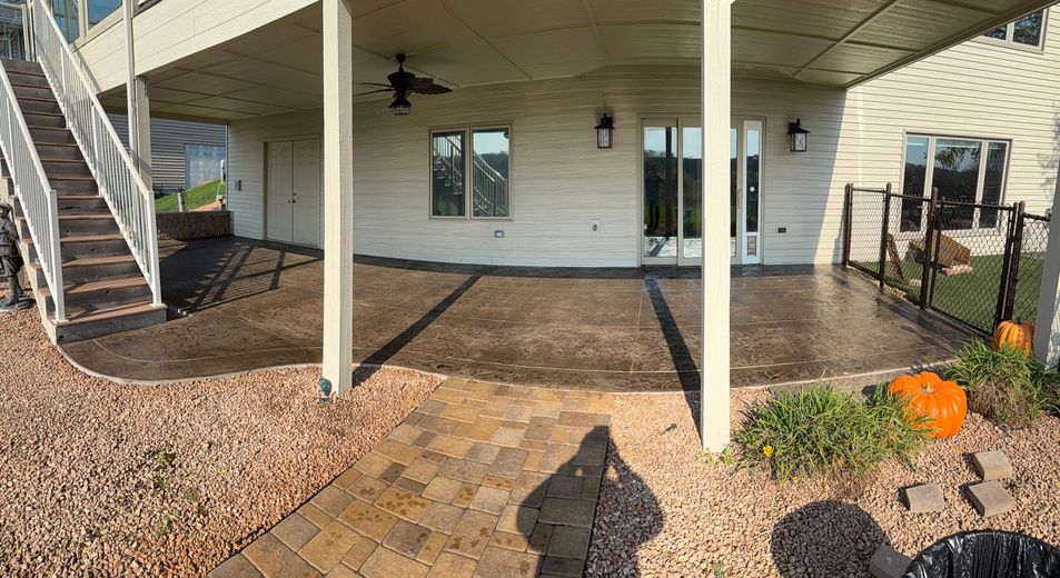 A patio under a house deck with stamped concrete flooring, a stone walkway, and a small decorative pumpkin on the grass.