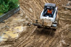 A person in an orange safety vest operates a skid-steer loader carrying dirt across a muddy construction site.