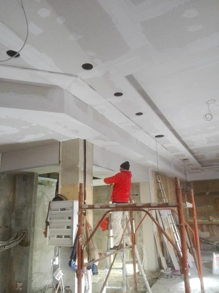 Un operaio con giacca rossa su un'impalcatura sta installando luci da soffitto in una stanza in costruzione.