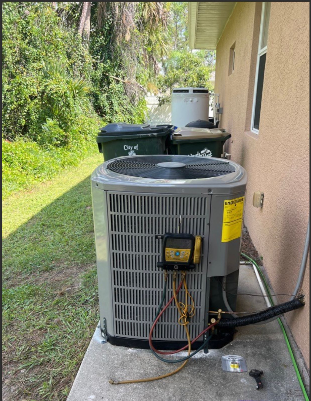 An air conditioning unit next to a building, with trash cans and foliage in the background.