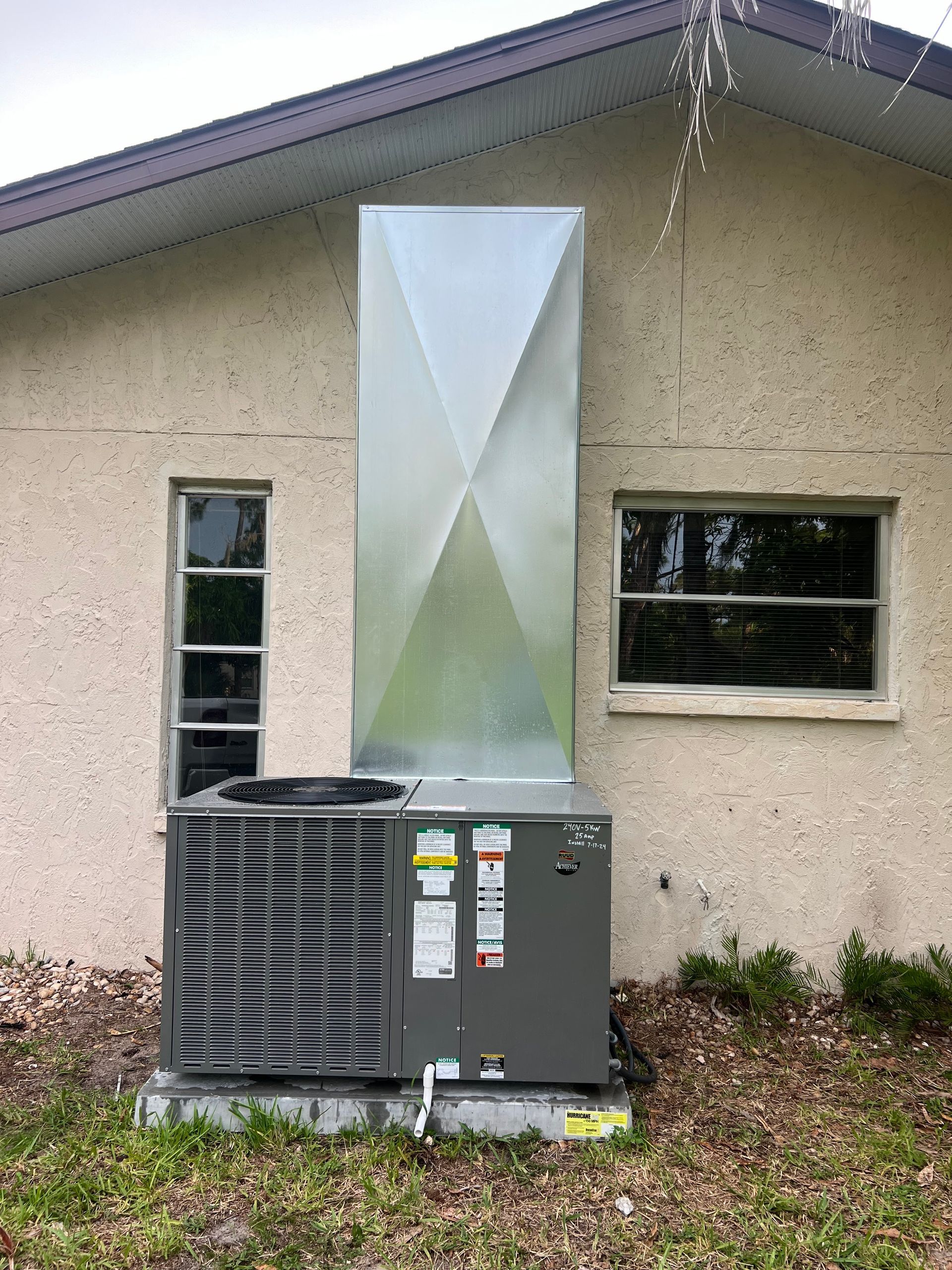 Outdoor air conditioning unit with metal ductwork, mounted against a light beige house wall, two windows, and greenery.