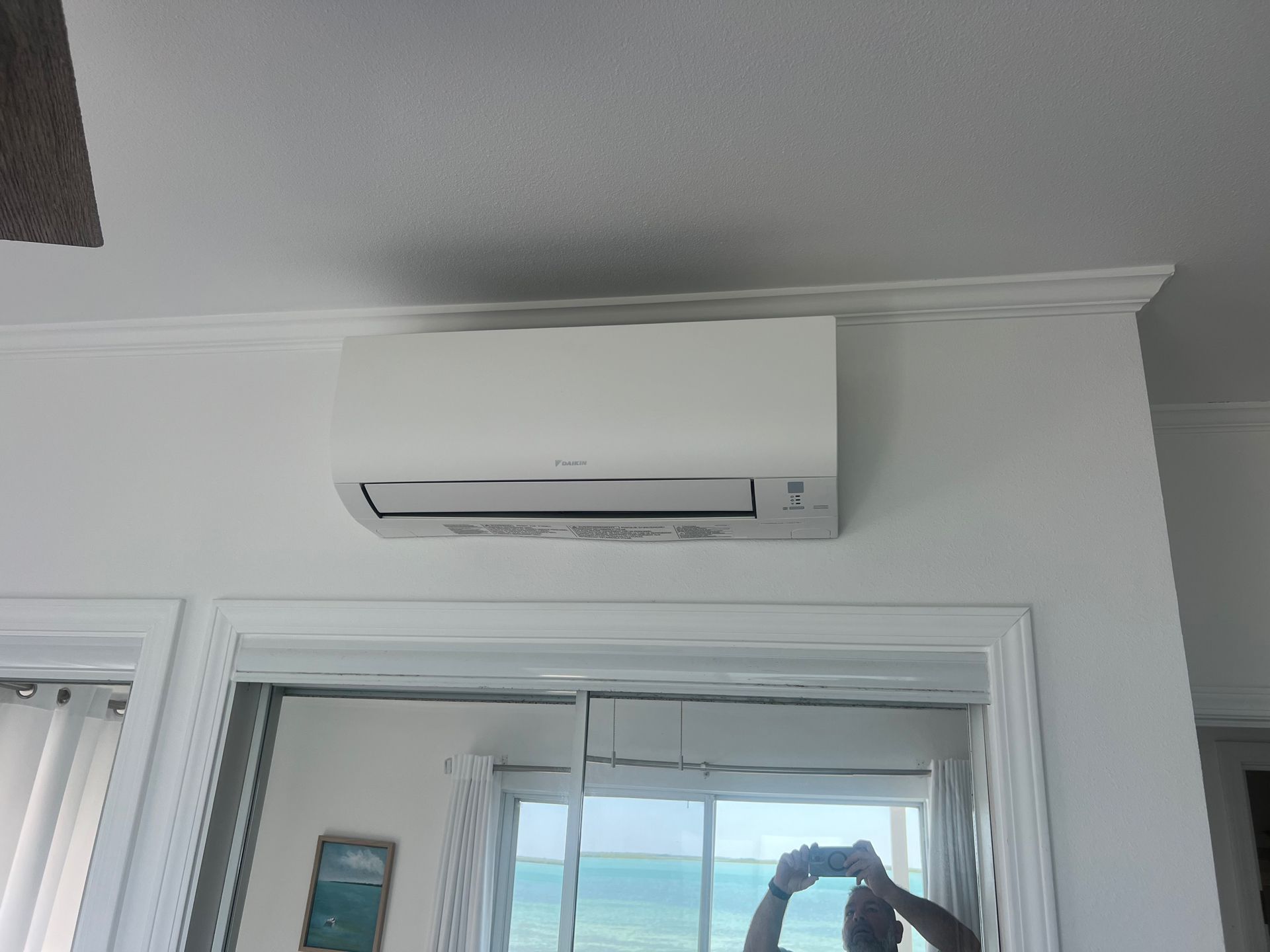 White air conditioner mounted on a white wall above a doorway with a mirror reflecting the view outside.
