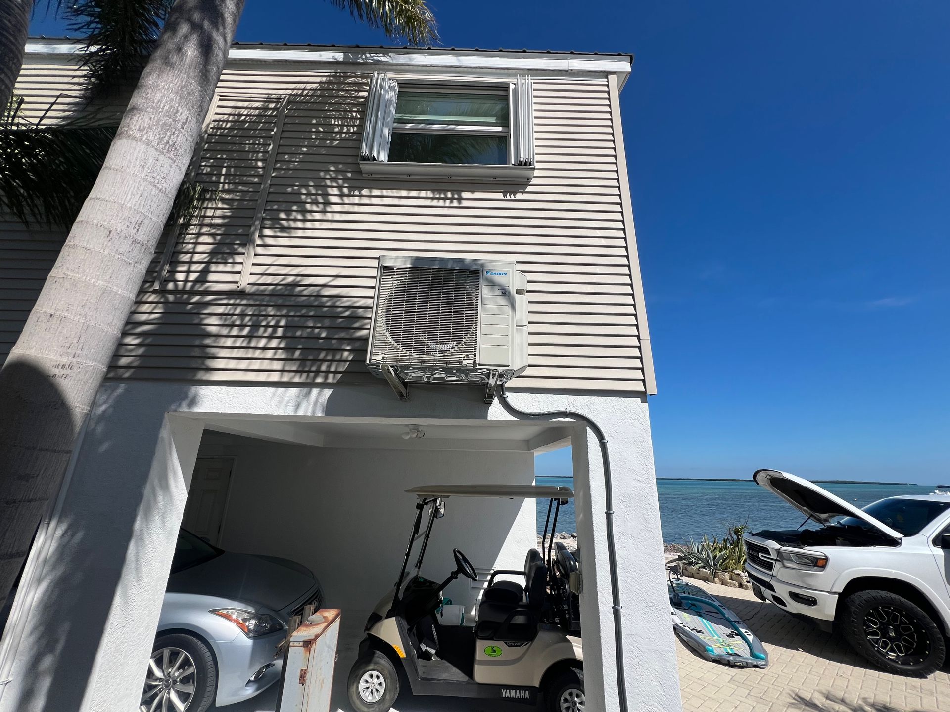 Two-story house with a garage. AC unit attached. Cars and golf cart parked in garage, surf board outside. Blue sky and ocean.