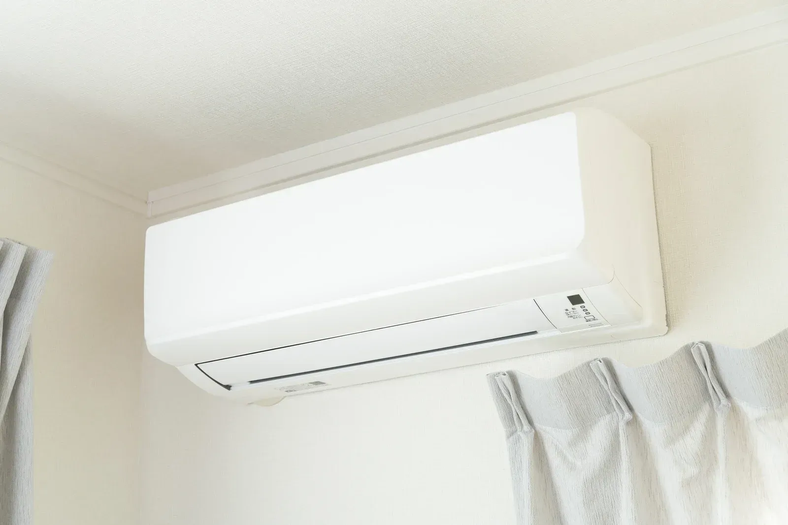 White wall-mounted air conditioning unit inside a room with white walls and gray curtains.