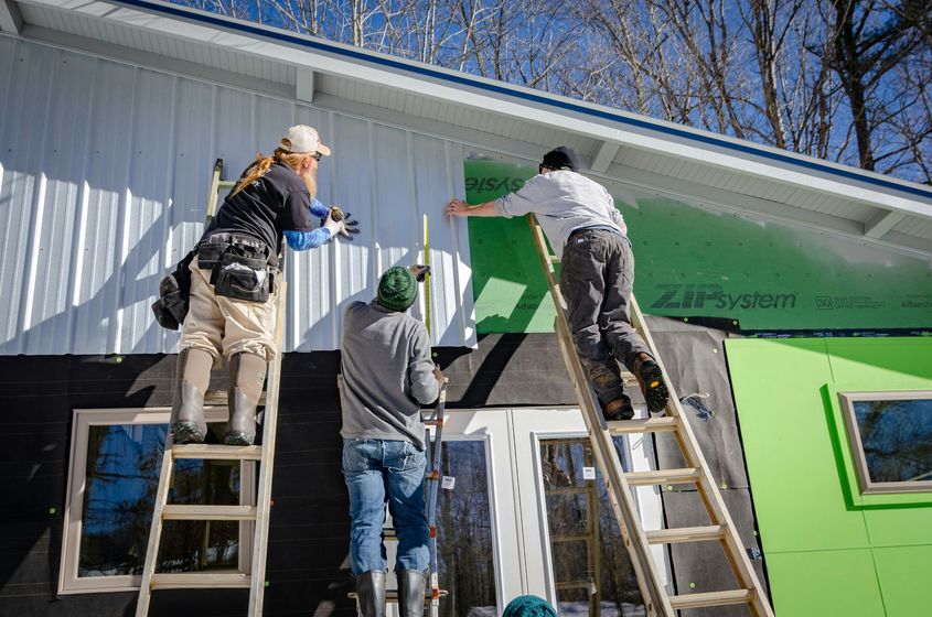 Three people on ladders install green house wrap on a building exterior.