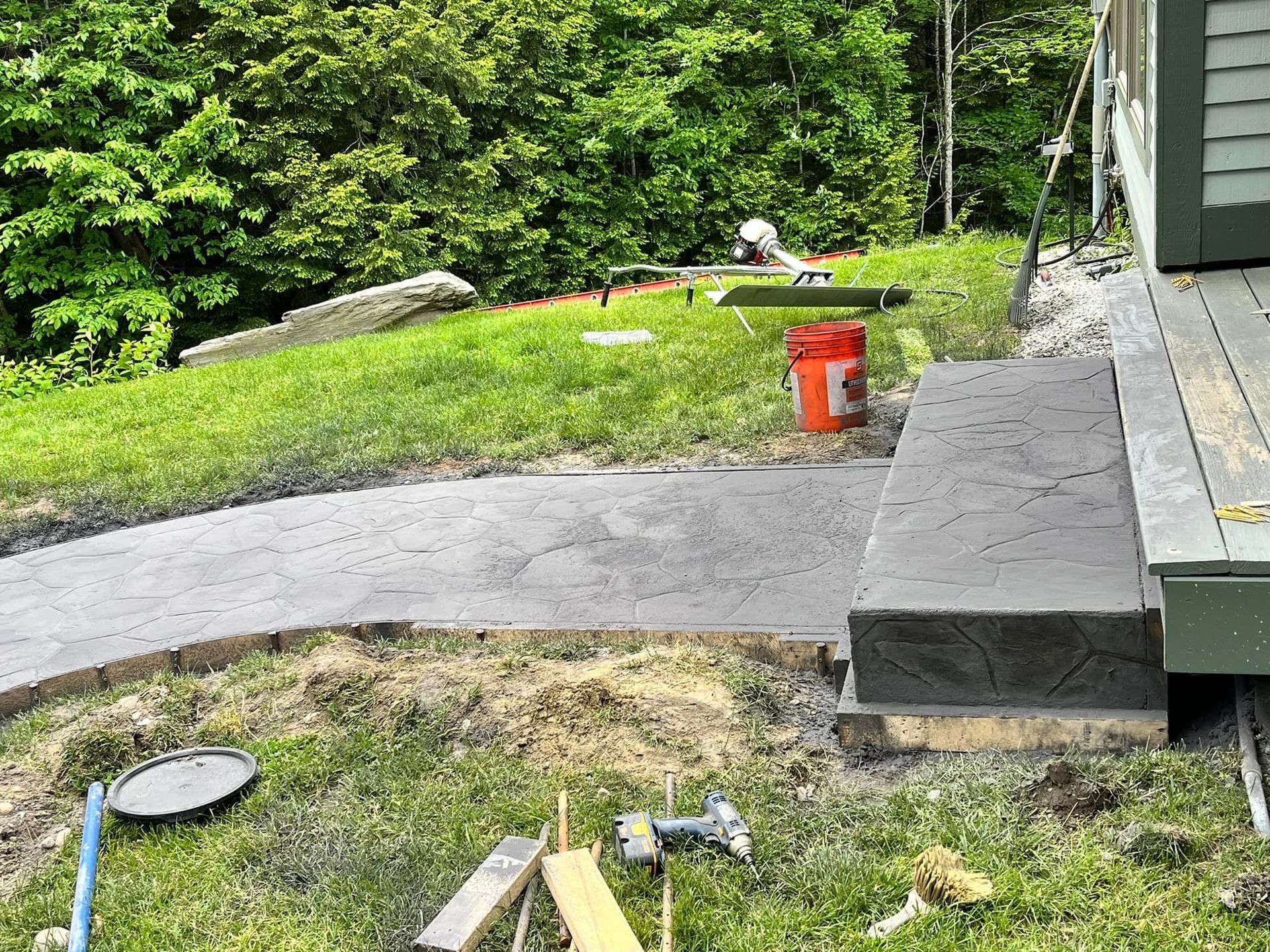 Backyard with a curved concrete path, grassy slope, orange utility bucket, and stacked pavers beside a house.