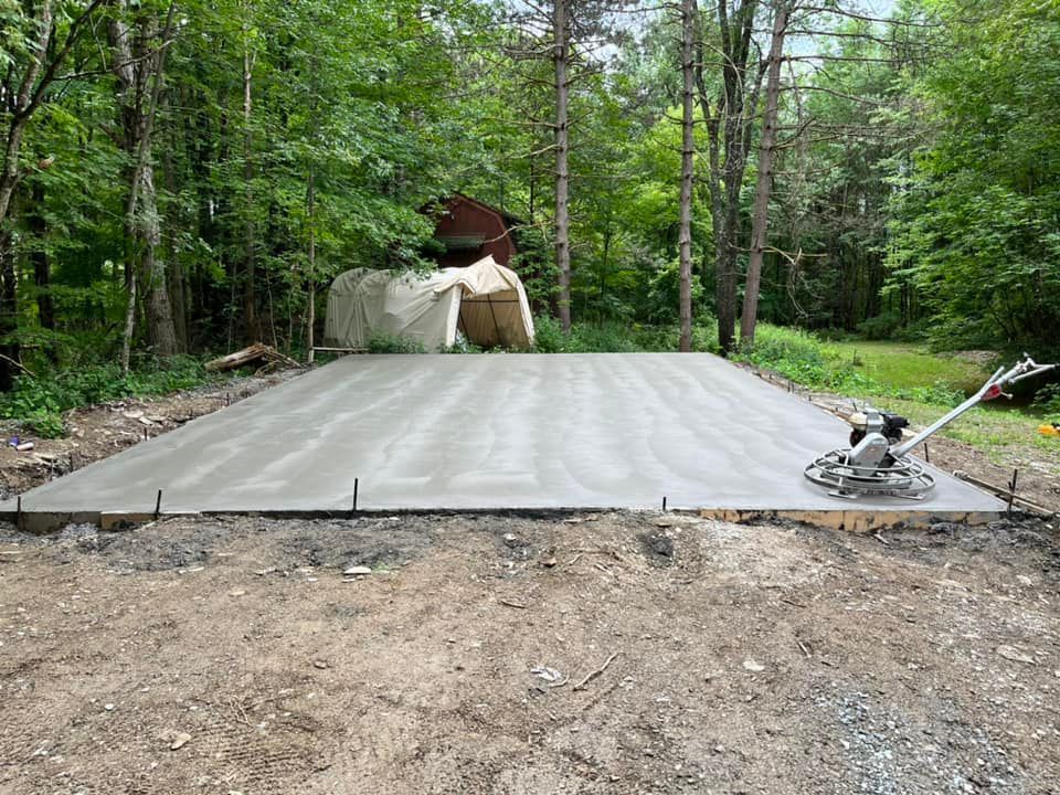 Freshly poured concrete slab in a wooded yard with a wheelbarrow and construction tools nearby