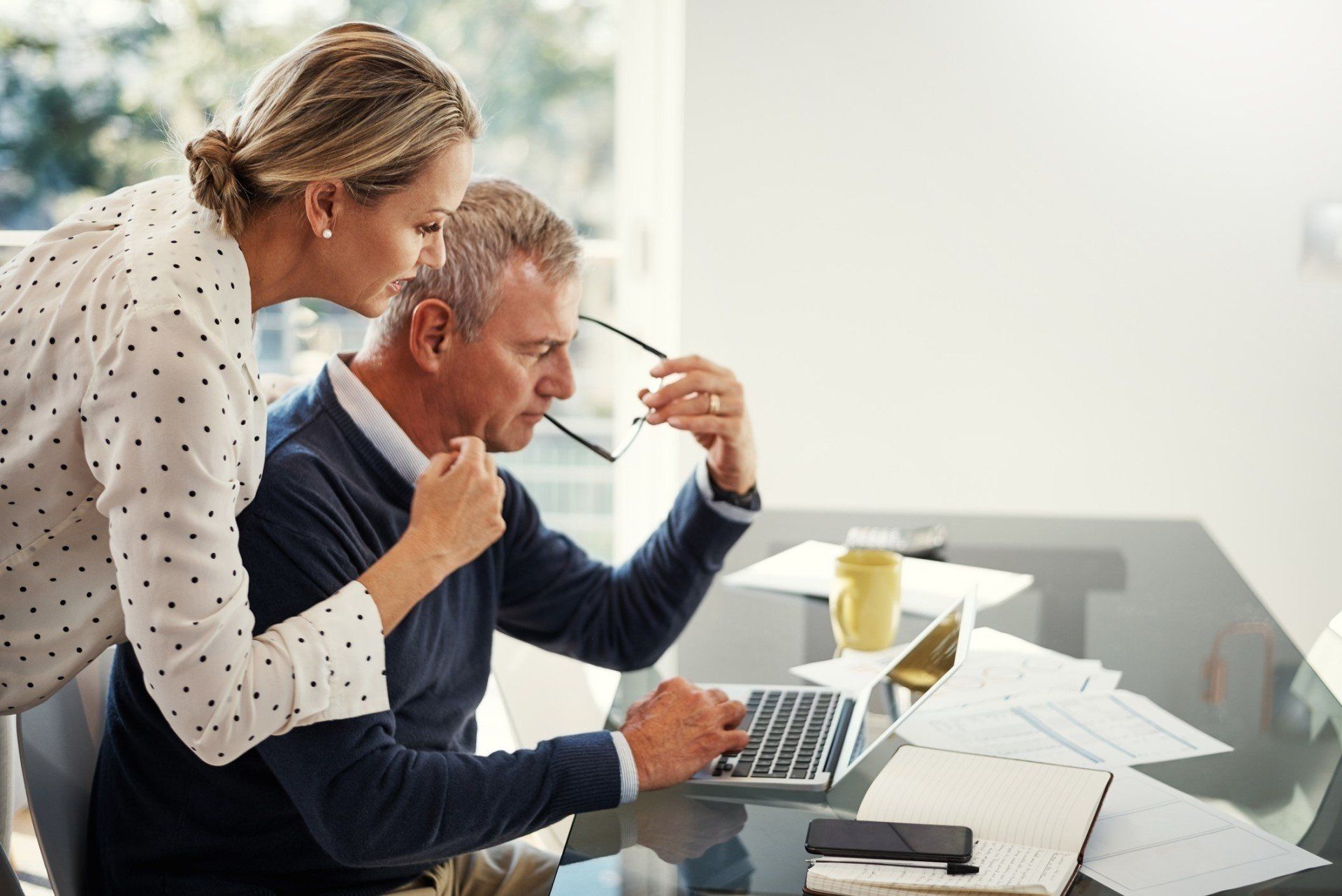 A woman is standing next to a man using a laptop computer.