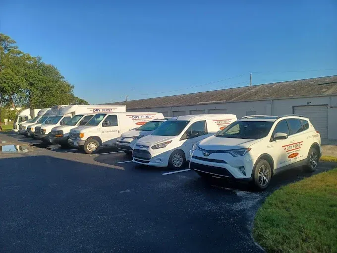 White vans and cars parked in a row, all with logo; sunny parking lot.