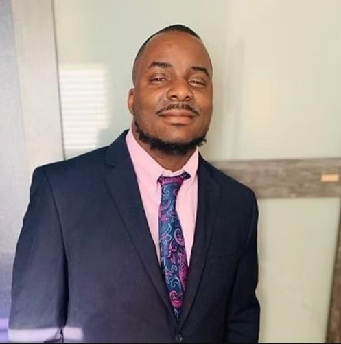 Man in a dark suit and pink shirt, wearing a patterned tie, smiling indoors.