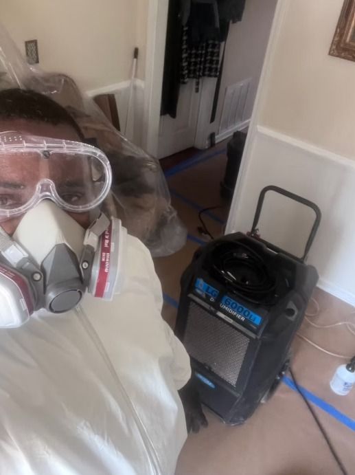 Person in protective gear near a dehumidifier in a room with blue tape on the floor.