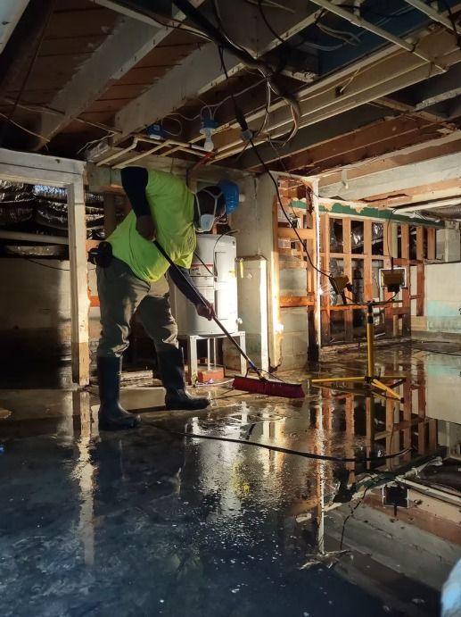A worker in protective gear sweeps water in a flooded basement under wooden beams and pipes.