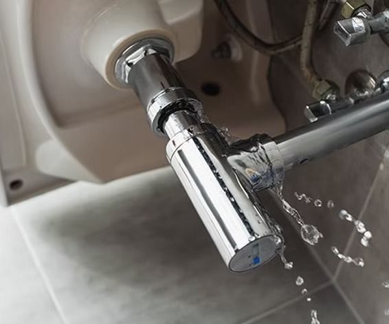 Leaking chrome plumbing under a white sink. Water drips from the pipe joint onto the floor.
