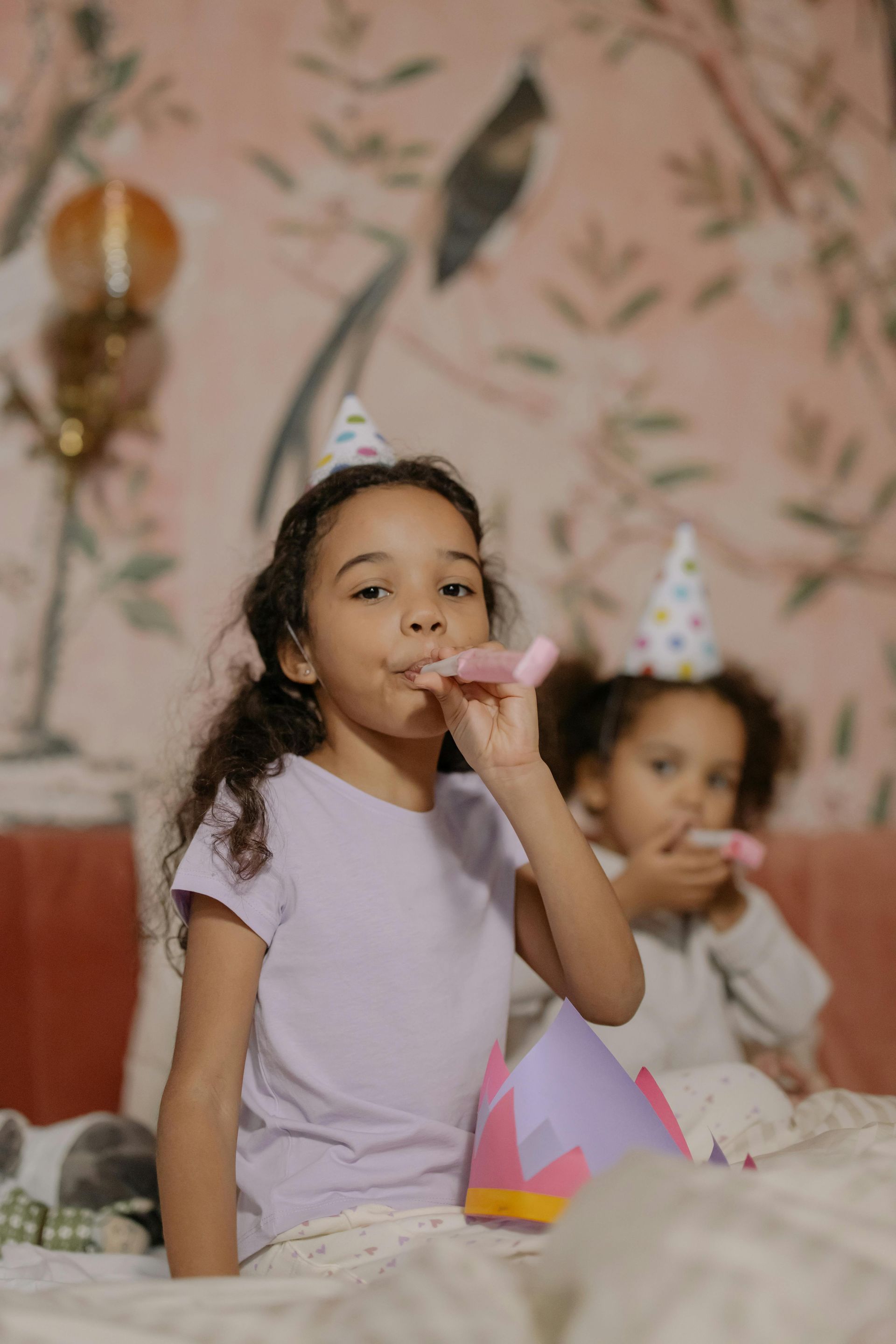 Due bambine sono sedute su un letto, indossano cappellini da festa e suonano le trombe da festa.