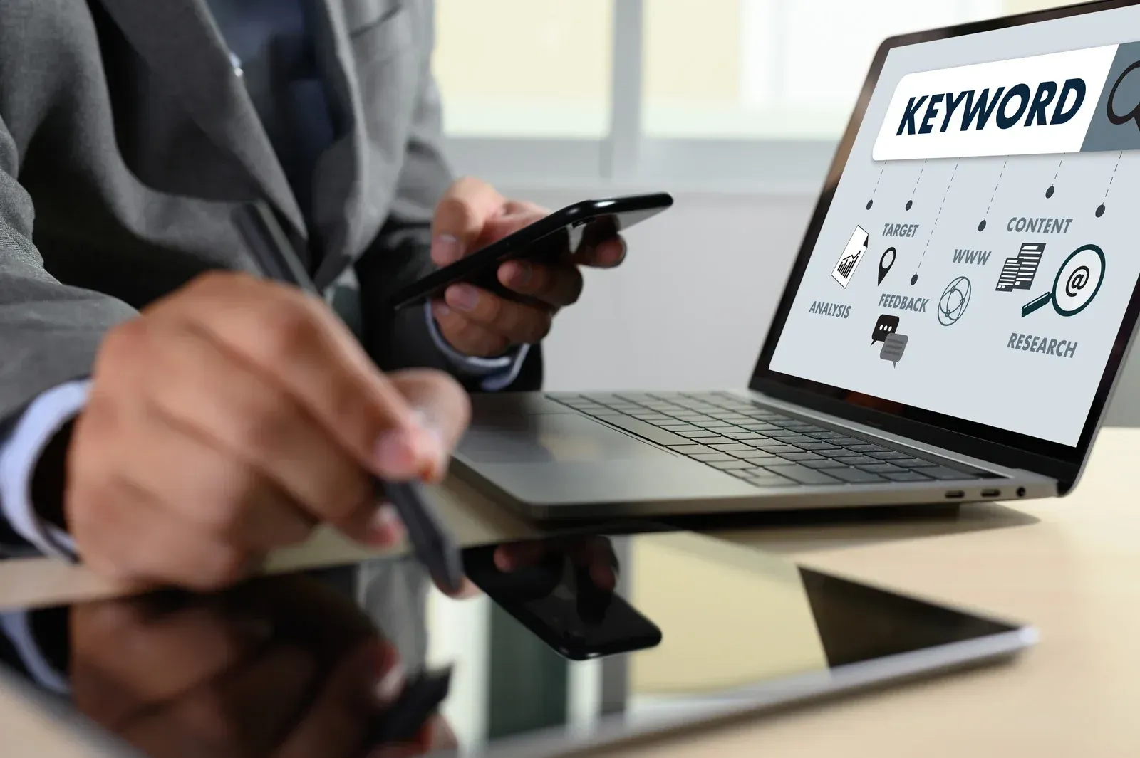 Person in suit using laptop, smartphone, and tablet for keyword research with a magnifying glass icon on screen.