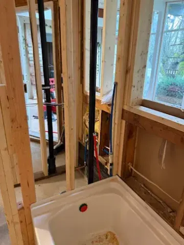 Unfinished bathroom framing with shower base, exposed studs, plumbing, and a window.