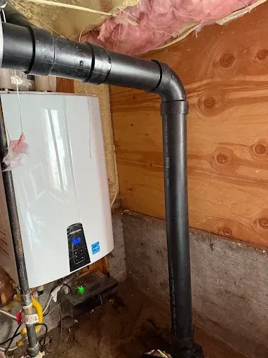Water heater and black vent piping in a basement with exposed wooden wall and insulation.