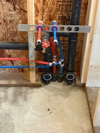 Exposed plumbing pipes and valves in unfinished wall framing, with white backer board and concrete floor.