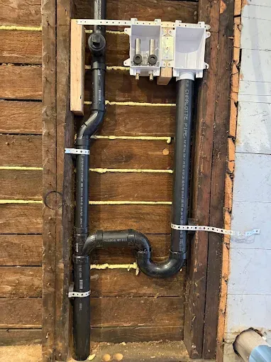 Black PVC plumbing pipes and a water meter mounted on an exposed wooden wall.