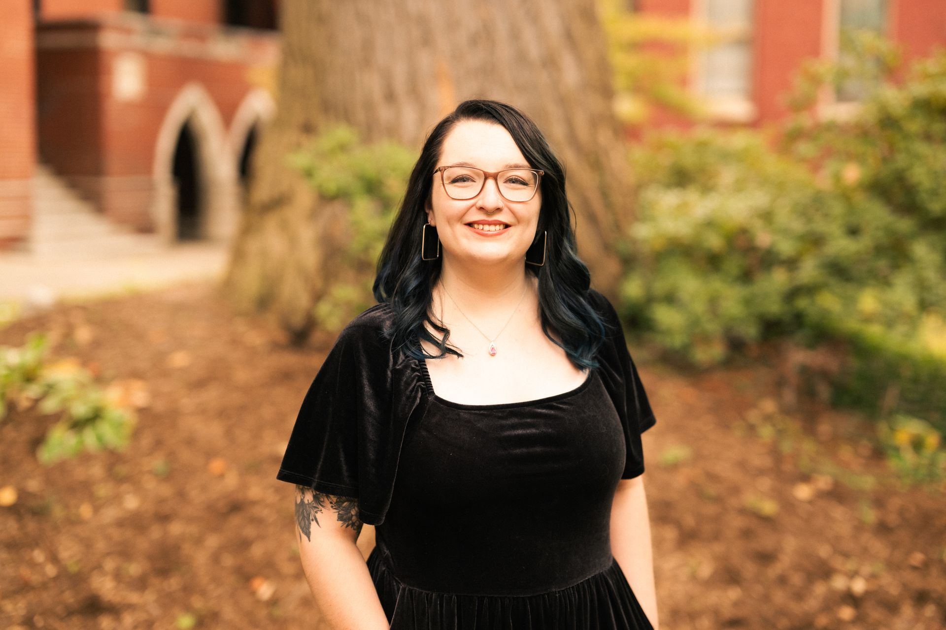 Woman with glasses smiles, wearing black dress and shrug, outdoors in front of tree and brick building.