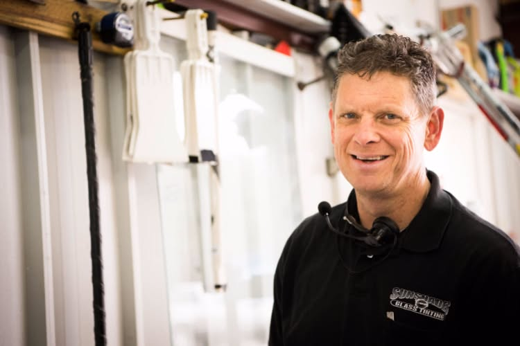 Man in a workshop smiling, wearing a black shirt with safety glasses, tools and equipment visible in the background.