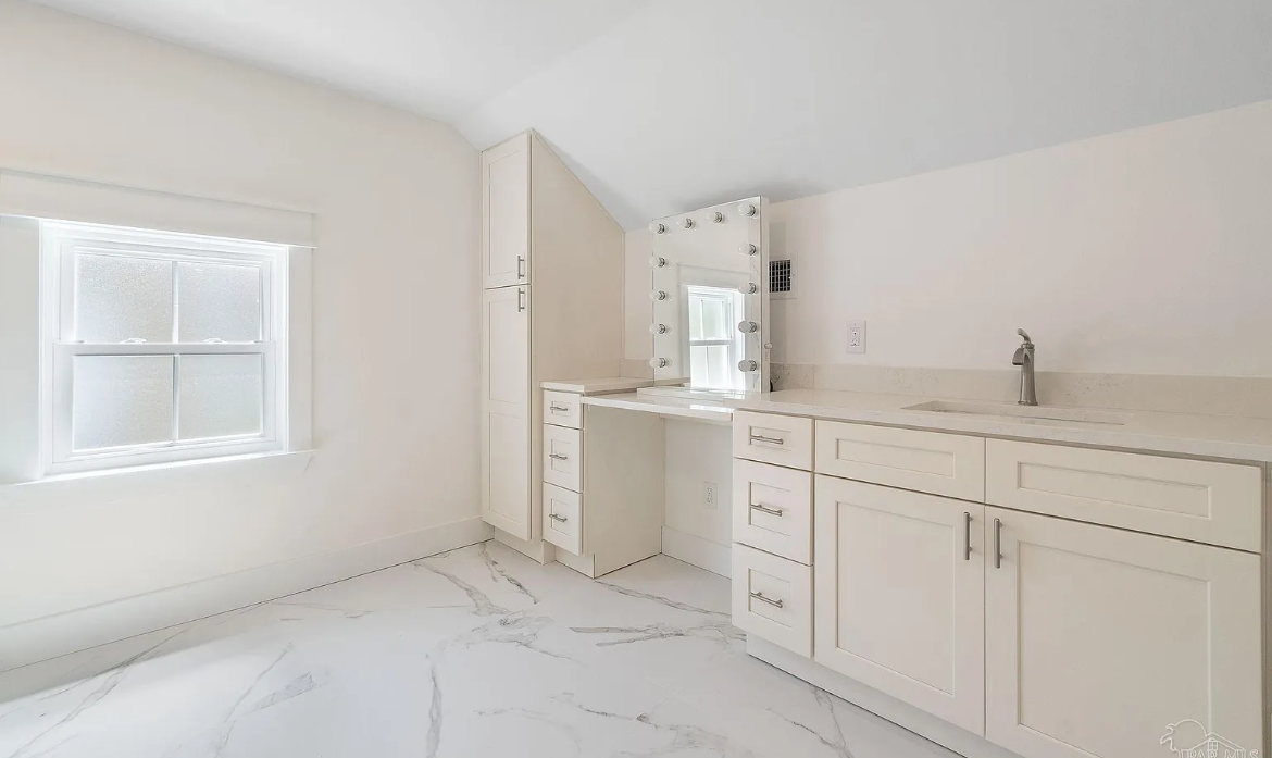 A bathroom with white cabinets , a sink , a mirror and a window.