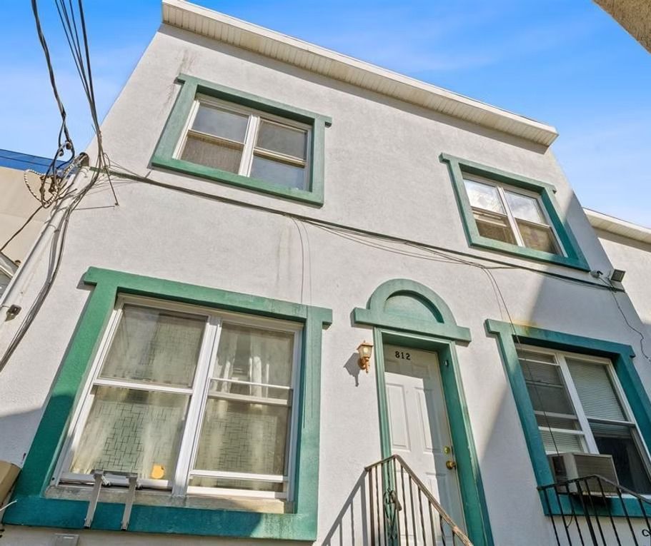 Two-story white building with green window trim, a white door, and overhead wires against a blue sky.