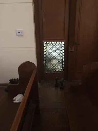 Wooden church pews face a confessional with a screened window; light streams in.