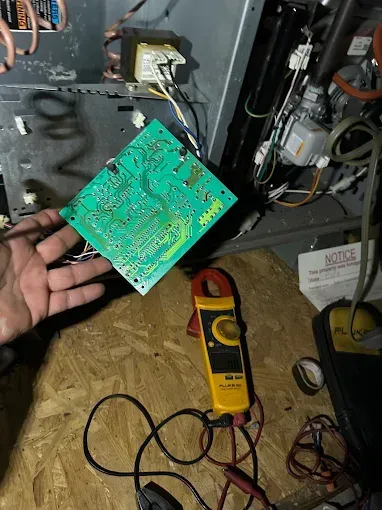 Hand holding a circuit board near a furnace. A multimeter lies on plywood.