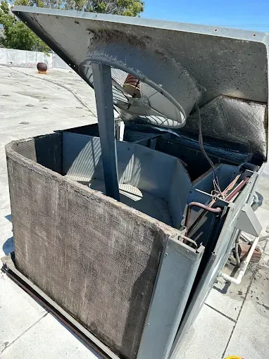 Rooftop air conditioning unit with open top; dirty coil and fan visible. Gray metal, outdoor setting.