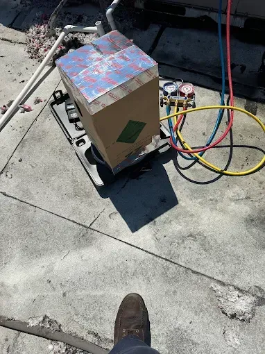A person's perspective, looking down at HVAC equipment on a concrete rooftop; a cardboard box with gauges and hoses connected.